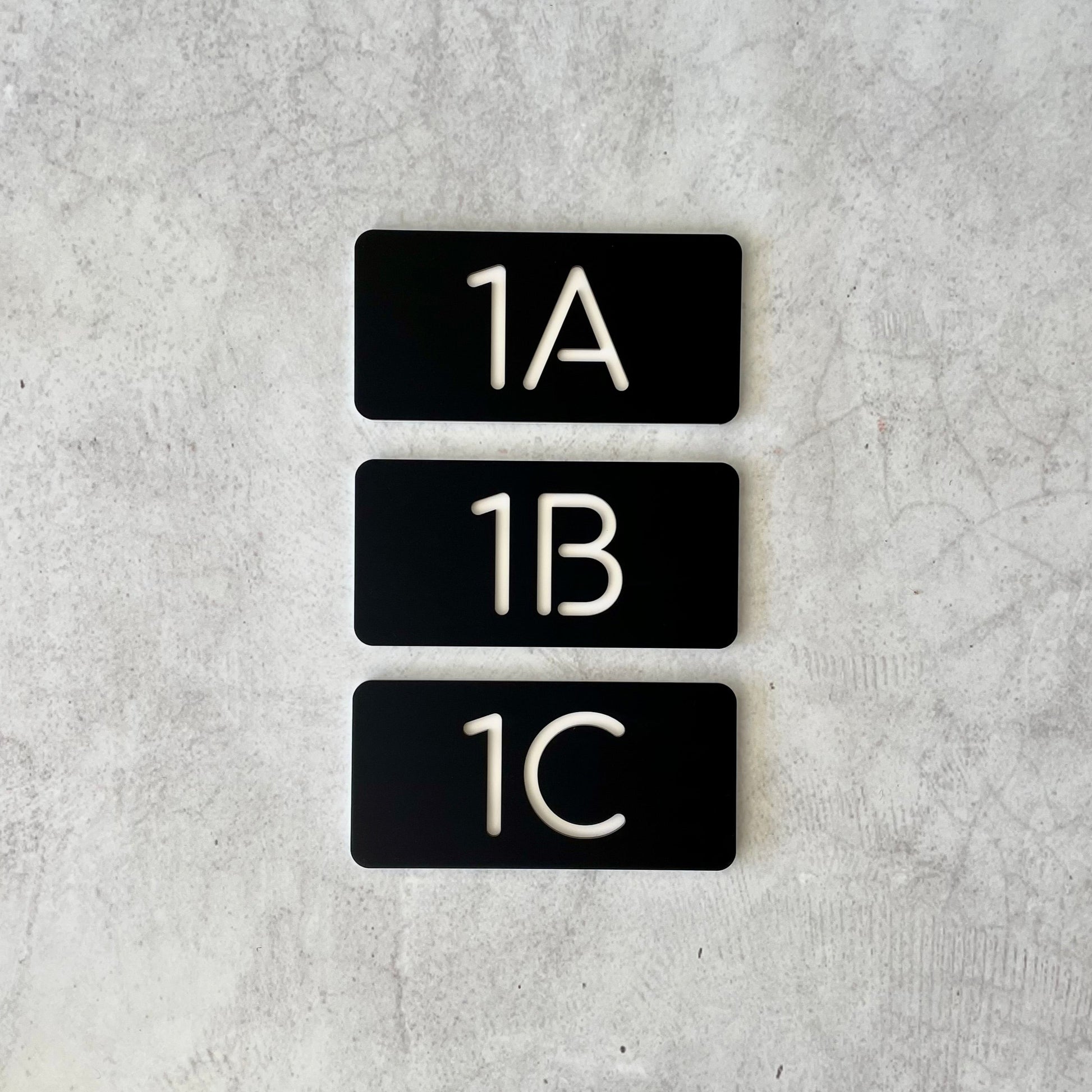 Three black rectangular house number signs with white text, arranged vertically, each with a different number: 1A, 1B, 1C.
