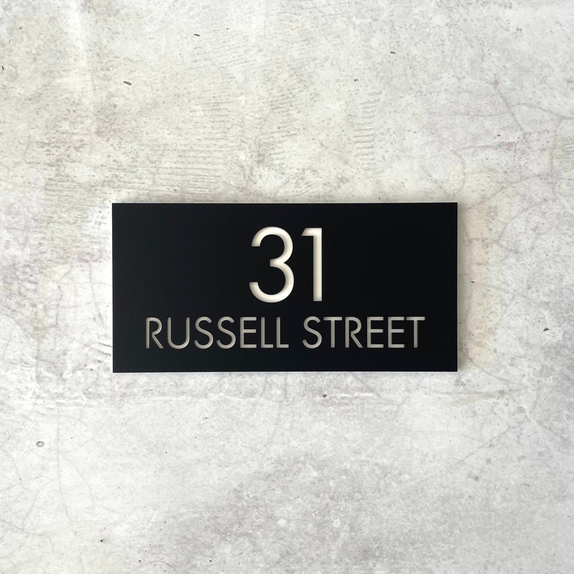 Black rectangular house number sign on a concrete wall.