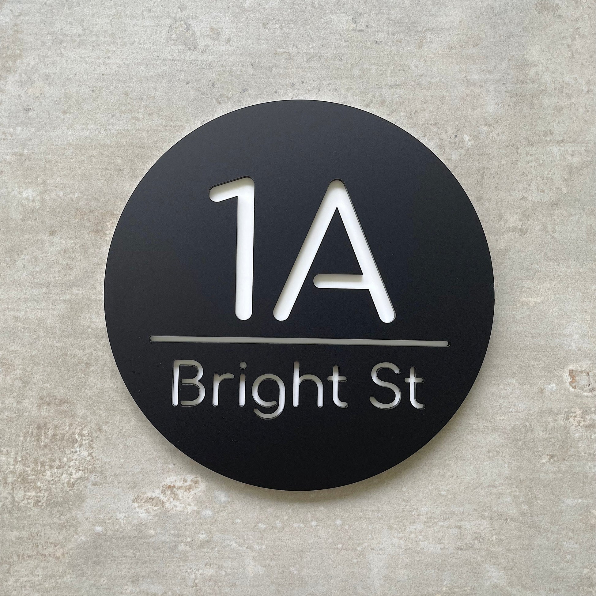 Round black house number sign with '1A Bright St' text on a textured gray background.