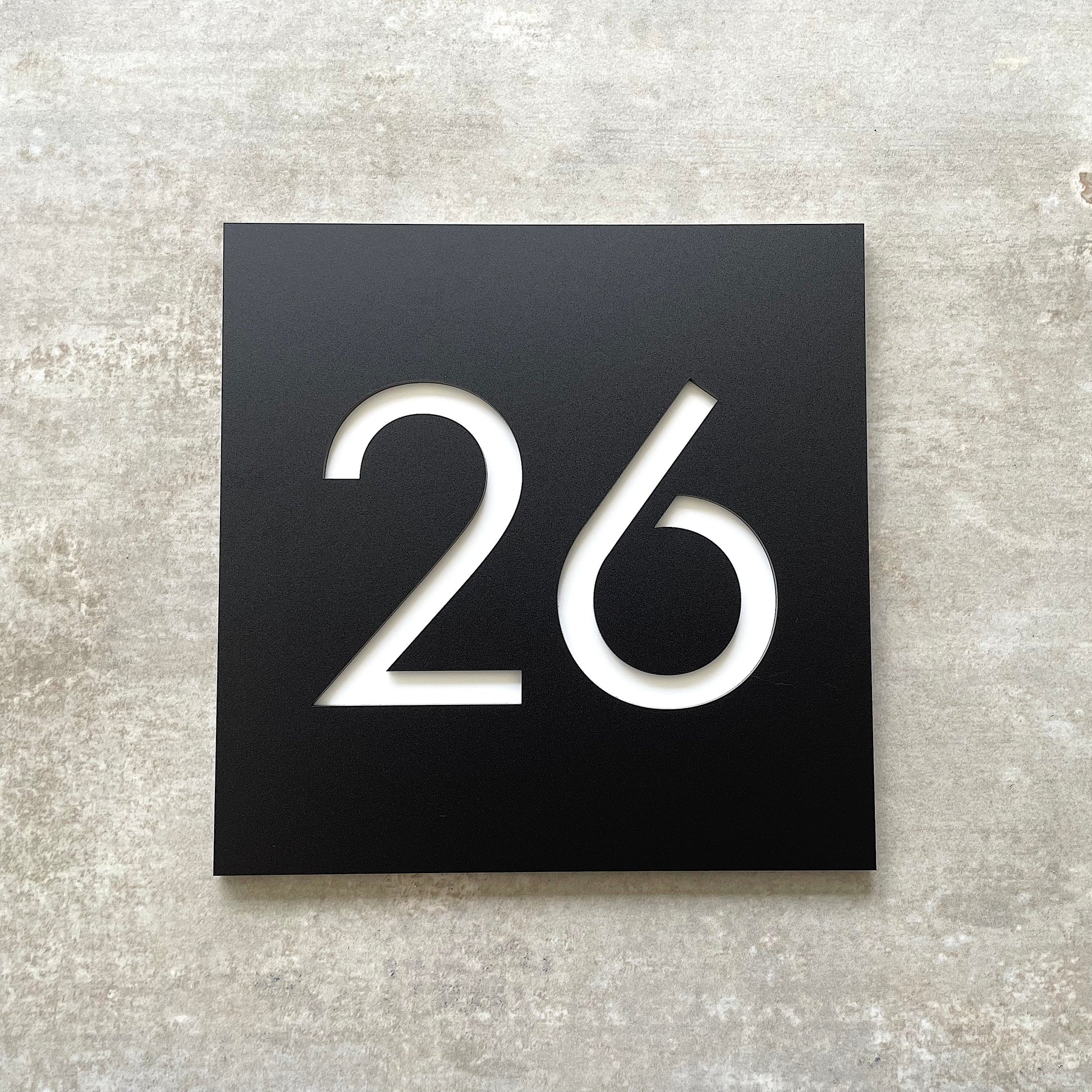 A square black house number sign with the number 26 in white.