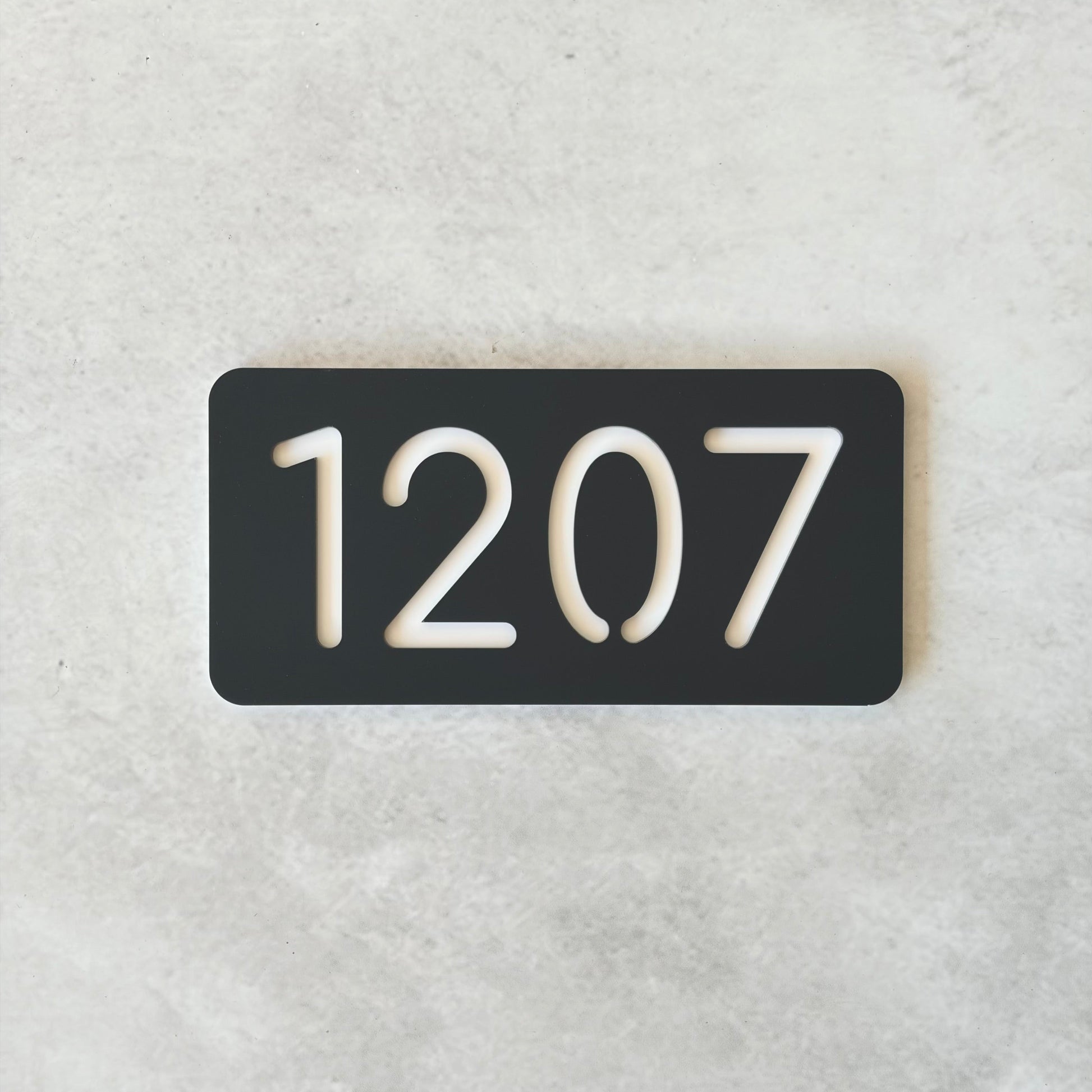 Charcoal rectangular plate with white apartment numbers '1207' on a light gray background