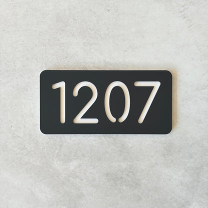 Charcoal rectangular plate with white apartment numbers '1207' on a light gray background
